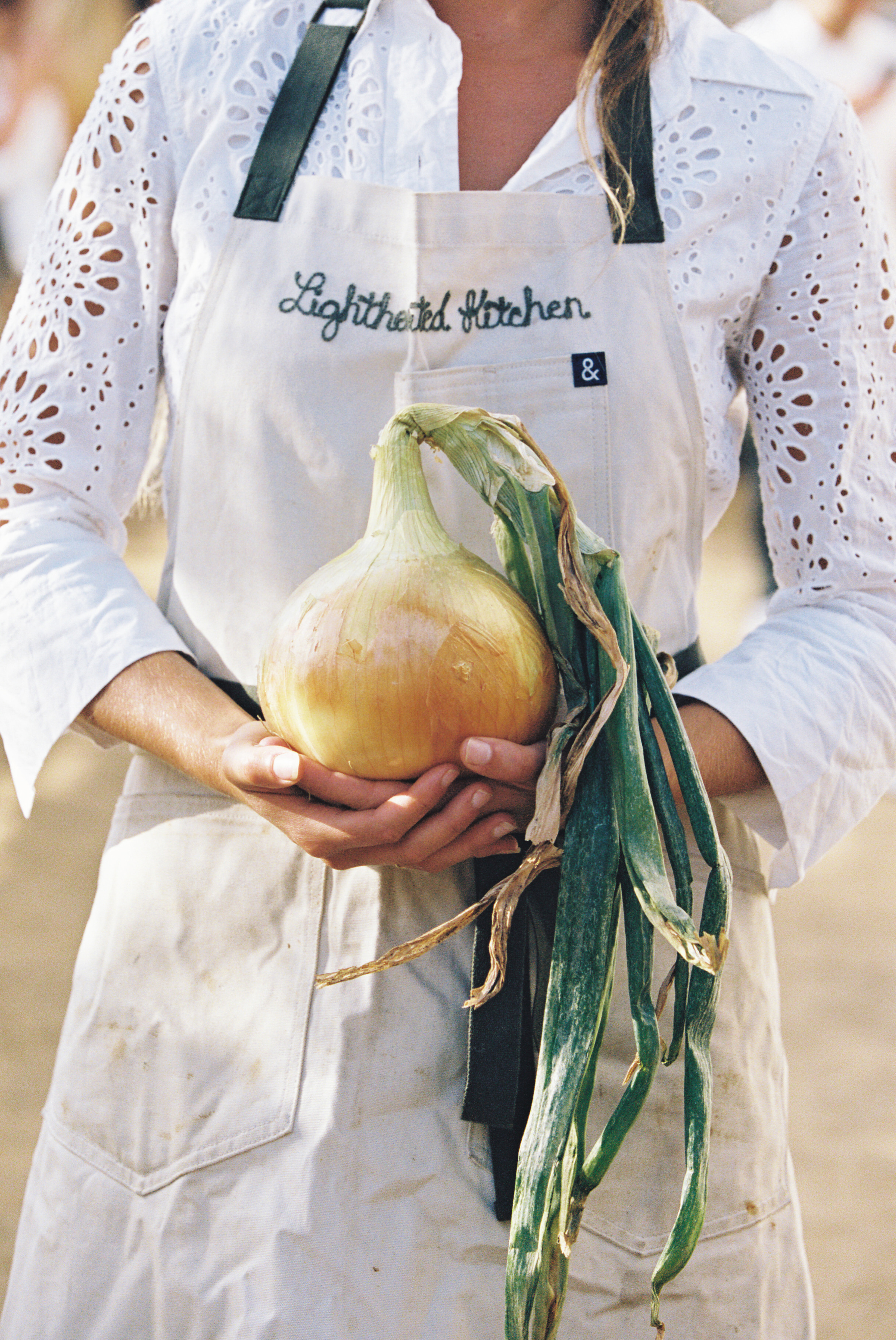 A Lighthearted Kitchen chef in an embroidered apron, holding a fresh-pulled onion with its greens still attached.